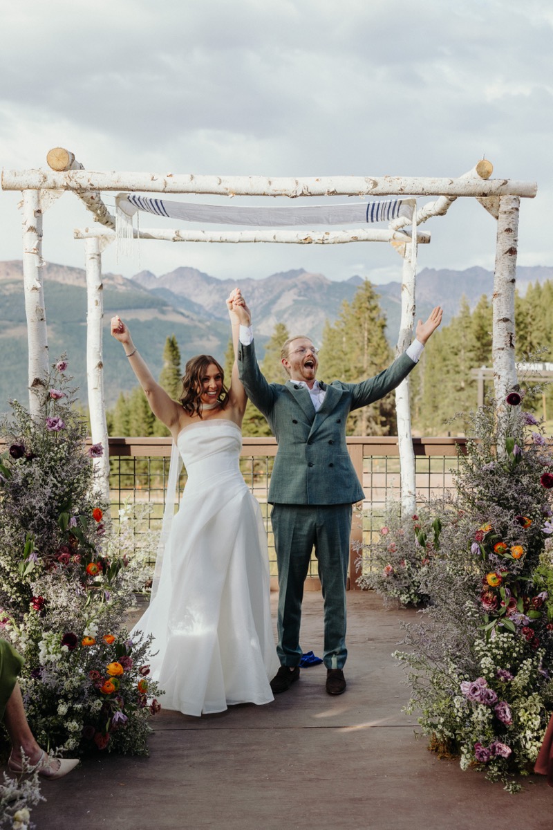 Jace and Amanda under a birch chuppah in the mountains, arms raised, just married.